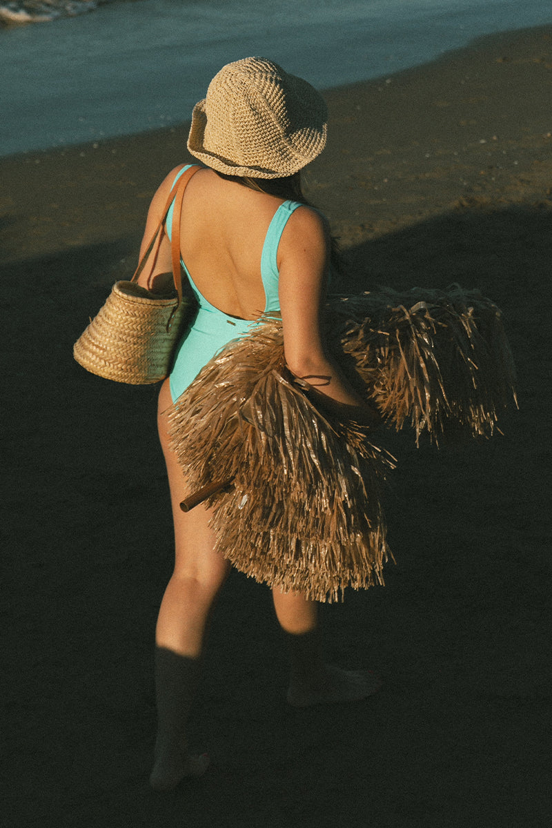 Model walks on the beach on her back wearing hat and blue AQVA Swimwear swimming costume